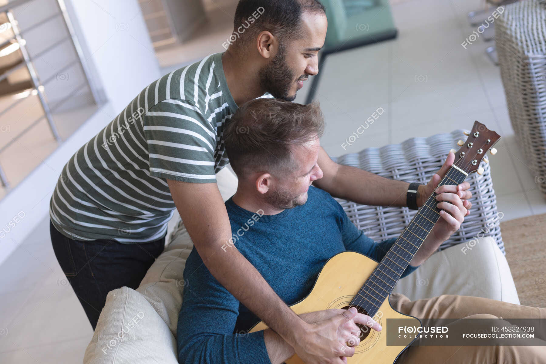 Multi Ethnic Gay Male Couple Sitting On Couch One Playing Guitar