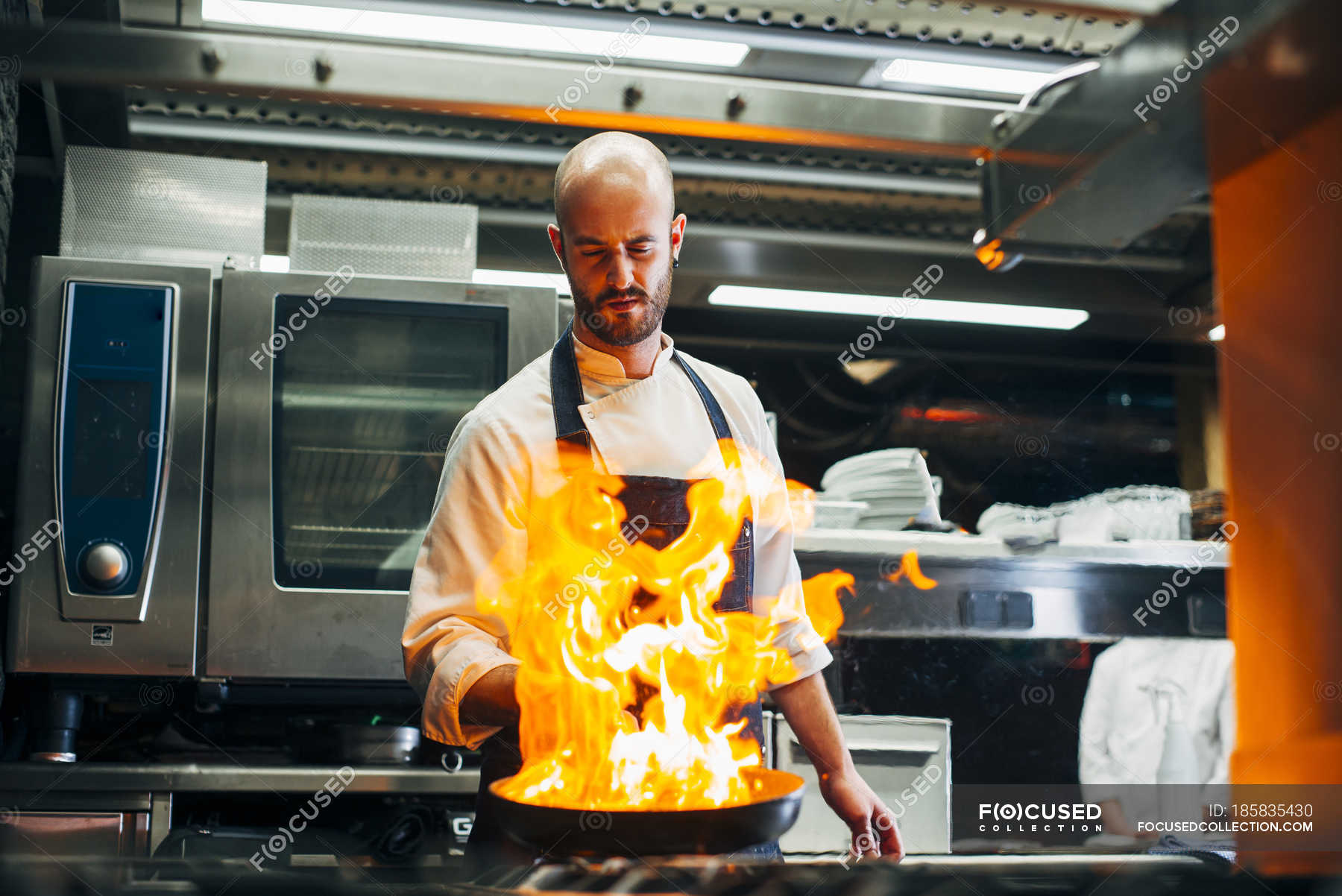 Chef Standing In Restaurant Kitchen And Making Flambe On Frying Pan