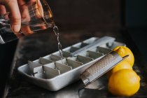 Mano de cultivo con vaso de agua llenando bandeja de hielo en el escritorio con rallador y limones - foto de stock