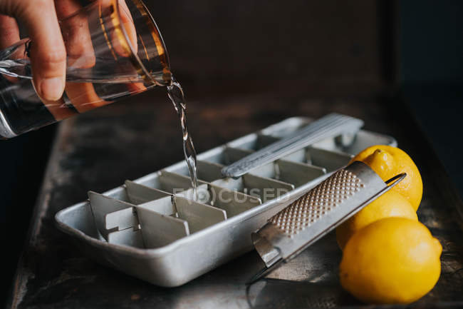 Рука для посева со стаканом воды, наполняющим поднос для льда на столе теркой и лимонами — стоковое фото
