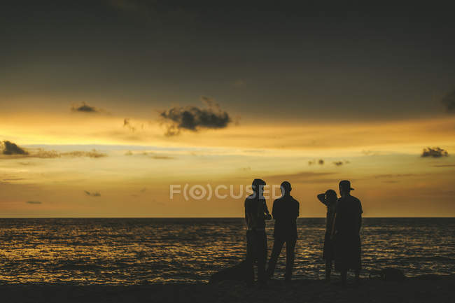 Люди стоять на піщаному березі з хвилястою водою над хмарним небом під час заходу сонця. — стокове фото