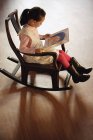 Chica sentada en silla y leyendo libro - foto de stock