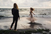 Вид сзади на молодого человека, смотрящего на веселую женщину, танцующую на песчаном пляже — стоковое фото