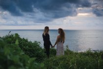 Вид сзади на молодую пару, держащуюся за руки и стоящую на приморском холме с закатом на заднем плане — стоковое фото