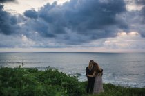 Rückansicht eines jungen Paares mit den Händen auf den Schultern, das auf einem Hügel am Meer steht und auf das Meer blickt — Stockfoto