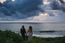 Vue arrière du jeune couple tenant la main et debout sur une colline au bord de la mer avec coucher de soleil en arrière-plan — Photo de stock