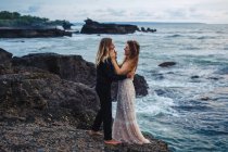 Vista lateral do casal romântico abraçando e amorosamente olhando um para o outro em rochas à beira-mar — Fotografia de Stock