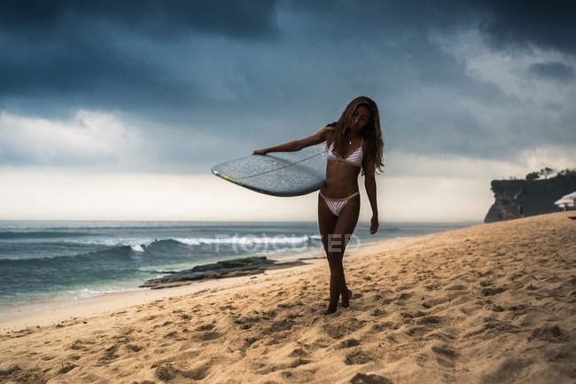 Жінка в гідрокостюмі, що несе дошку для серфінгу на пляжі — стокове фото