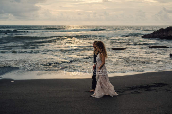 Giovane coppia che cammina sulla spiaggia sabbiosa e si tiene per mano al tramonto — Foto stock