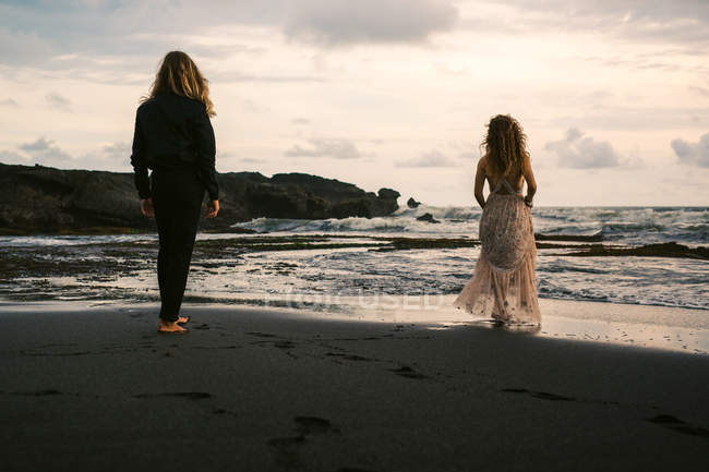 Vista trasera del joven hombre y la mujer caminando en la playa de arena al atardecer - foto de stock