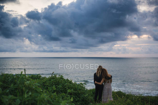 Rückansicht eines jungen Paares mit den Händen auf den Schultern, das auf einem Hügel am Meer steht und auf das Meer blickt — Stockfoto