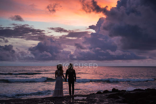 Rückansicht eines jungen Paares, das sich an den Händen hält und am Strand steht, mit Sonnenuntergang im Hintergrund — Stockfoto
