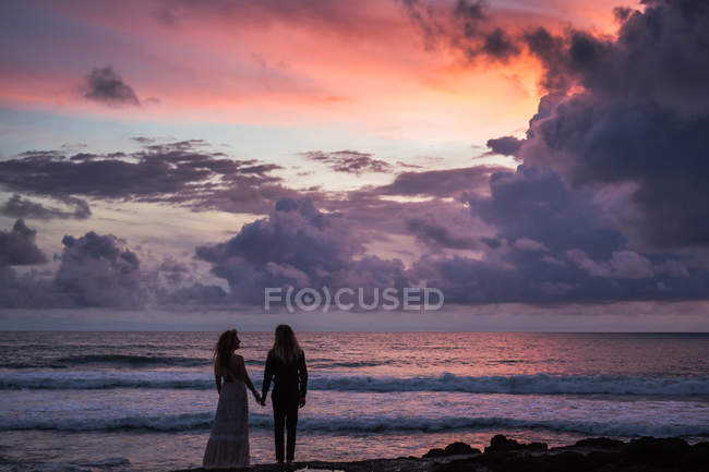Rückansicht eines jungen Paares, das sich an den Händen hält und am Strand steht, mit Sonnenuntergang im Hintergrund — Stockfoto