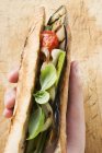 Hand holding grilled vegetables and basil in baguette over wooden surface — Stock Photo