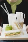 Primo piano vista ritagliata della donna che tiene panini, mela e bevande sul vassoio — Foto stock