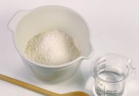 Primo piano vista di farina in ciotola e acqua in brocca di misura su fondo bianco con cucchiaio di legno — Foto stock