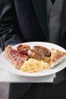 Englisches Frühstück: Speck, Ei, Wurst und Bohnen, auf weißem Teller in der Hand, Mittelteil — Stockfoto