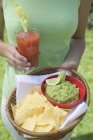 Daytime cropped view of woman holding tomato drink and basket of guacamole and chips — Stock Photo