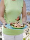 Обрезанный вид женщины с тарелкой крабовых когтей, фигуркой танцовщицы и соусом на пляже — стоковое фото