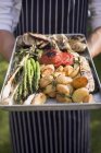 Man in apron  holding aluminium tray of grilled vegetables in hands. midsection — Stock Photo