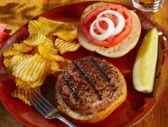 Открытый гамбургер с помидорами, луком, чипсами и огурцом на тарелке с вилкой — стоковое фото