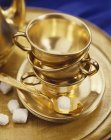 Closeup view of golden tea cups, saucers and spoon with sugar cubes — Stock Photo
