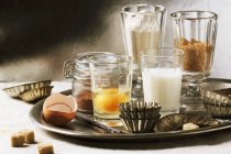 Vários ingredientes de cozimento em copos, incluindo farinha, ovo, açúcar mascavo, leite e latas de bolo antiquadas na bandeja — Fotografia de Stock