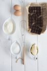 Top view of flour with eggs, whisk, butter, sugar and cracked chocolate bar — Stock Photo