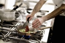 Шеф-кухар додає нарізані помідори на сковороду з ножем в руках — стокове фото