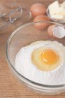 Вид крупным планом сырого яйца поверх муки в стеклянной миске, рядом с яйцами, маслом и венчиками — стоковое фото