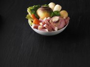 A bowl of fresh vegetables with bacon on black wooden surface — Stock Photo