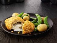 Chicken Kiev with mange tout and potatoes on black plate over table — Stock Photo
