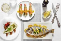 Три разных блюда из рыбы с лимонами и белым вином на белых тарелках за столом — стоковое фото