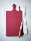 Top view of a red wooden chopping board and a knife on a tea towel — Stock Photo