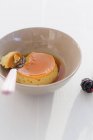 Closeup view of Creme caramel with spoon in bowl and one blackberry on white surface — Stock Photo