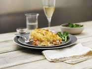 Mixed fish pie with broccoli and wine on black plates over table — Stock Photo