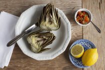 A boiled artichoke with tomato salsa and lemon on white plate with knife over wooden surface — Stock Photo