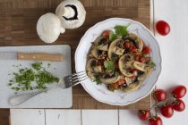 Cogumelos fritos com tomates e salsa em placa branca sobre mesa de madeira com garfo e faca — Fotografia de Stock