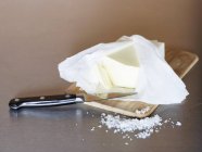 Vue rapprochée du beurre dans un emballage ouvert sur une planche de bois avec du sel — Photo de stock