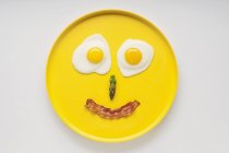Cara sonriente en una placa amarilla hecha de dos huevos fritos, espárragos y una tira de tocino - foto de stock