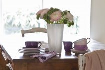 Un tavolo in legno con stoviglie impilate e un mazzo decorativo di fiori e foglie di ranuncolo — Foto stock