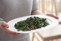 Person holding dish with blanched spinach with raisins and pine nuts — Stock Photo
