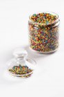 Closeup view of colored sugar sprinkles in glass jar on white surface — Stock Photo