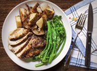 Нарезанный жареный цыпленок, брокколини и жареная картошка с тимьяном на белой тарелке, установленной на деревянном столе с голубой клетчатой салфеткой — стоковое фото