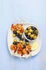 Brochettes de crevettes et d'oliviers et olives marinées sur plaque blanche sur surface bleue — Photo de stock