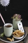 Savoury snacks with mug of tea and stem of flowers on black wooden board against dark background — Stock Photo