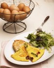 Омлет с жареными овощами на белой тарелке с вилкой и сырыми яйцами на заднем плане — стоковое фото