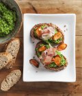 Crostini topped with pea cream, roasted carrot slices and ham  on white plate over wooden surface — Stock Photo
