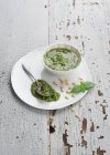 Closeup view of Pesto in a white bowl and on a plate with pine nuts, spoon and leaves — Stock Photo