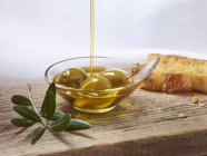 Aceite de oliva que se vierte sobre aceitunas verdes en un plato pequeño sobre la superficie de madera - foto de stock